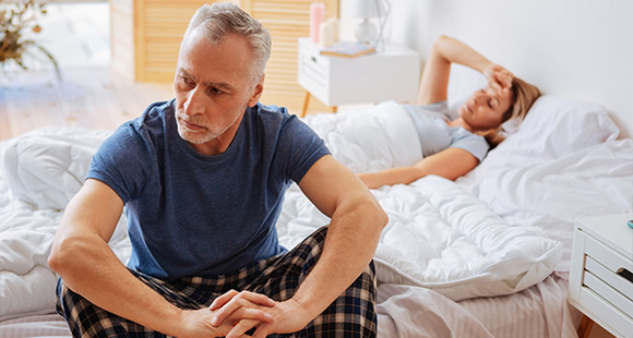 an older man sitting on the edge of a bed with a woman laying on the other side